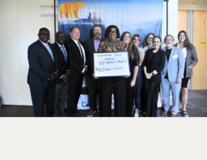 A group of people standing together indoors in front of an Employment Training Panel banner. One person in the center holds a whiteboard that reads “September Panel 2026, 66 Approved Projects, $28.3 Million Funded.” The group is posed for a photo and standing in a well-lit room with doors and signage visible in the background.