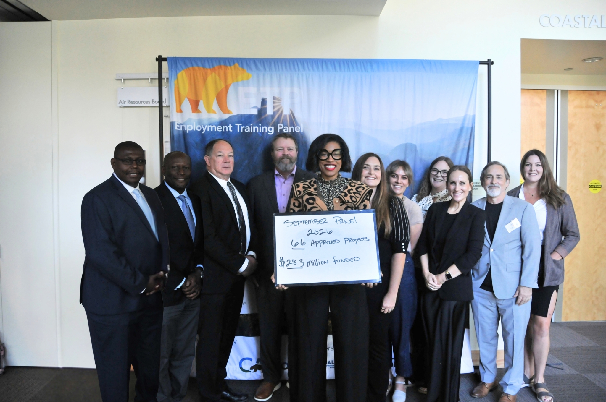 A group of people standing together indoors in front of an Employment Training Panel banner. One person in the center holds a whiteboard that reads “September Panel 2026, 66 Approved Projects, .3 Million Funded.” The group is posed for a photo and standing in a well-lit room with doors and signage visible in the background.